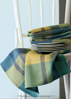 A collection of blue and green checkered dish towels, showcasing intricate patterns made using 4-shaft overshot techniques, is neatly folded on a chair. Some of the towels hang over the backrest, partially veiling a set of white plates beneath. These designs are inspired by projects featured in "Best of Handwoven, Top Ten Projects in Overshot Vol III," an eBook by Long Thread Media.