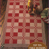 A wooden table is adorned with a handwoven table runner in a red and beige checkered pattern, exemplifying exceptional craftsmanship on four shafts. Nesting dolls rest on the runner, complemented by a plate featuring a red and white design. Text reads, "Long Thread Media: Best of Handwoven, Top Ten Table Runners on Four Shafts".