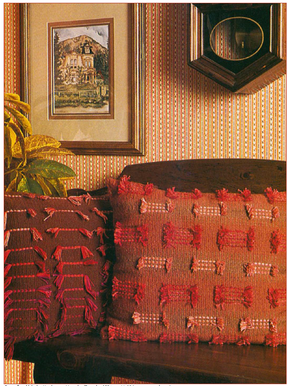 Two decorative pillows with red and pink tassels, crafted using techniques highlighted in the "Best of Handwoven New Technique Series: Rigid Heddle Pattern Book 1" by Long Thread Media, rest on a dark wooden bench. A plant is positioned to the left, and a painting of a building adorns the striped wallpapered wall. An octagonal mirror hangs above the pillows.