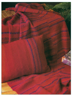 A wooden chair is adorned with a red blanket featuring blue stripes, woven using the techniques from the "Best of Handwoven New Technique Series: Rigid Heddle Pattern Book 1" by Long Thread Media. A matching red striped cushion complements the ensemble. In the background, a green fern introduces a natural element, and a floral-patterned box is stationed on a nearby wooden surface.