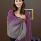 A woman with long dark hair is smiling while wrapped in a cozy purple and grey striped shawl. She is standing inside against a brown wall, with a framed picture in the background, possibly taking inspiration from the patterns in her copy of "Best of Handwoven, Rigid Heddle Technique & Pattern Book 3" by Long Thread Media.