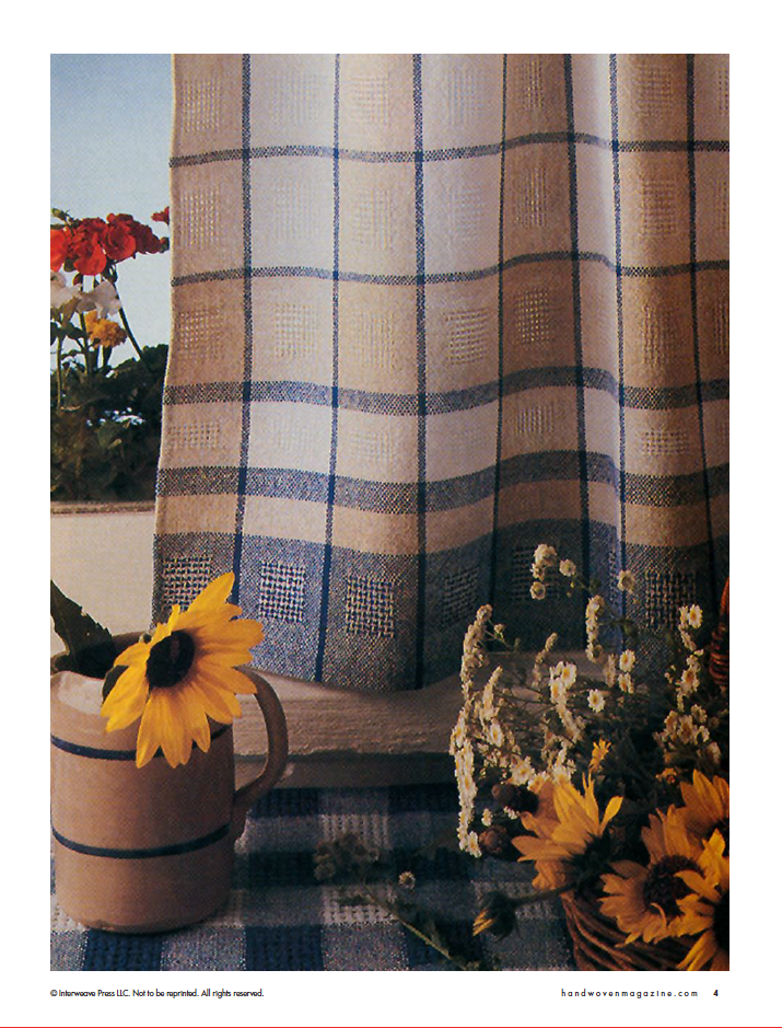 In front of a plaid-patterned curtain, the "Best of Handwoven, Reader's Choice: Top Ten Projects from 30 Years of Handwoven eBook (Printed version)" by Long Thread Media creates a cozy scene with sunflowers artfully arranged in a ceramic jug and vase. A checkered cloth adds a rustic flair underneath, while red flowers softly emerge on the left to enhance the warm atmosphere of these beautifully woven household textiles.