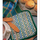 A cozy household scene features intricate weaving techniques, represented by the *Best of Handwoven* from Long Thread Media, alongside wooden butter paddles. Nearby, a white cup containing a hot beverage sits beside freshly baked biscuits on a metal tray. In the background, a colorful tartan cloth adds an extra touch of charm.