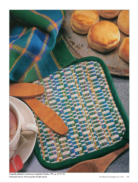 A cozy household scene features intricate weaving techniques, represented by the *Best of Handwoven* from Long Thread Media, alongside wooden butter paddles. Nearby, a white cup containing a hot beverage sits beside freshly baked biscuits on a metal tray. In the background, a colorful tartan cloth adds an extra touch of charm.