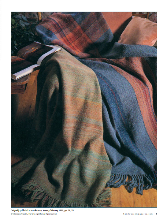 A cozy armchair draped with two fringed, multicolored wool blankets beautifully exhibits the craftsmanship of doubleweave. Resting on the chair's armrest is a copy of "Best of Handwoven, Doubleweave, Doublewidth!" by Long Thread Media. The scene is set against a partially visible wooden floor, creating a warm and inviting atmosphere.