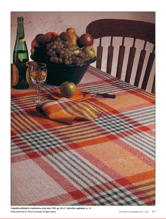 The dining table features a vibrant plaid tablecloth from the "Best of Handwoven, Doubleweave, Doublewidth!" collection by Long Thread Media. It holds a green bottle of wine, a glass of white wine, an apple, and a bowl brimming with fruit such as grapes, pears, and bananas. A wooden chair is partially visible beside the table.