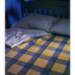 A neatly made bed with a wooden headboard features two pillows and a folded, striped shirt. The bedspread, echoing the style found in the "Best of Handwoven, Doubleweave, Doublewidth!" eBook by Long Thread Media, displays a plaid pattern in yellow, gray, and blue tones that creates a cozy atmosphere.