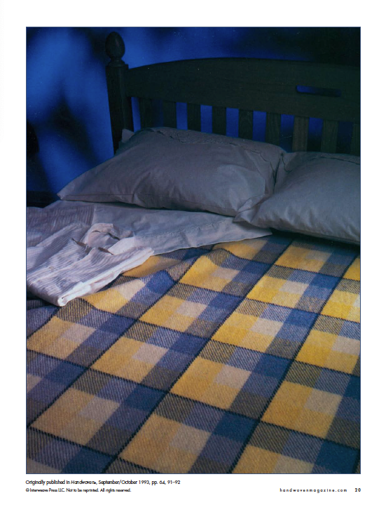 A neatly made bed with a wooden headboard features two pillows and a folded, striped shirt. The bedspread, echoing the style found in the "Best of Handwoven, Doubleweave, Doublewidth!" eBook by Long Thread Media, displays a plaid pattern in yellow, gray, and blue tones that creates a cozy atmosphere.