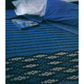 The bed is elegantly covered with a handwoven blue bedspread featuring dark and light diagonal stripes. A magazine from Long Thread Media's "Best of Handwoven, Doubleweave, Doublewidth!" eBook (Printed version) rests on the intricate doubleweave fabric, while in the background, a white pillow stands out against a blue pillowcase.