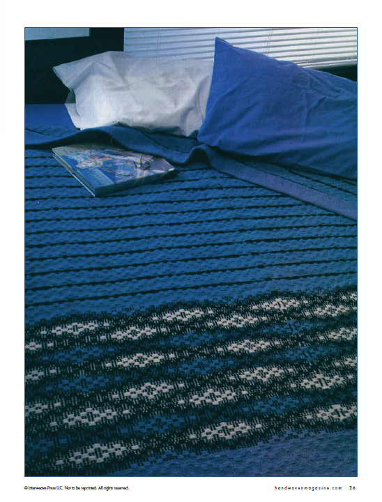 The bed is elegantly covered with a handwoven blue bedspread featuring dark and light diagonal stripes. A magazine from Long Thread Media's "Best of Handwoven, Doubleweave, Doublewidth!" eBook (Printed version) rests on the intricate doubleweave fabric, while in the background, a white pillow stands out against a blue pillowcase.