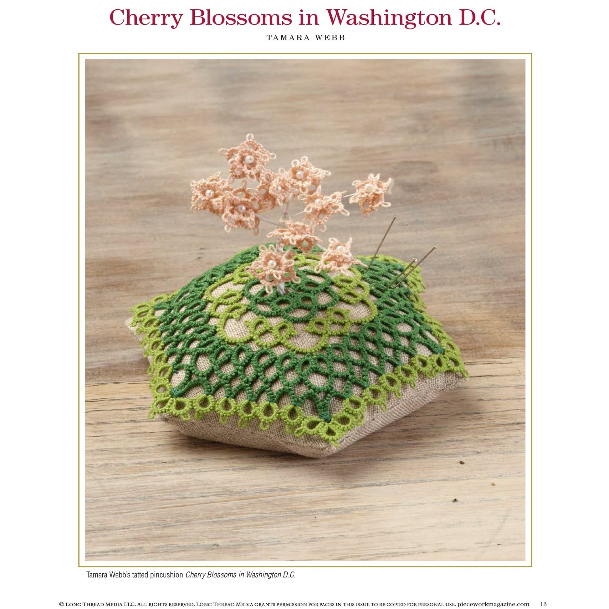 A cushioned pincushion adorned with a green lace cover showcases delicate, tatted pink cherry blossoms emerging from its top. It rests on a wooden surface, under the title *"Cherry Blossoms in Washington D.C."* and created by Tamara Webb. This beautiful piece is featured in the eBook, *PieceWork Presents: Pincushions 5 New Pincushion Designs to Bead, Crochet, Tat, Cross-Stitch, and Appliqué*, published by Long Thread Media.