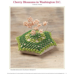 A cushioned pincushion adorned with a green lace cover showcases delicate, tatted pink cherry blossoms emerging from its top. It rests on a wooden surface, under the title *"Cherry Blossoms in Washington D.C."* and created by Tamara Webb. This beautiful piece is featured in the eBook, *PieceWork Presents: Pincushions 5 New Pincushion Designs to Bead, Crochet, Tat, Cross-Stitch, and Appliqué*, published by Long Thread Media.