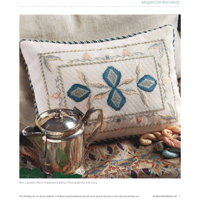 A silver teapot sits next to an intricately embroidered pillow featuring blue and beige patterns. The pillow showcases symmetrical designs with a touch of elegance. Nearby, 'Elegant Embroidery with 5 Beautiful Projects to Stitch' by Long Thread Media is open, revealing colourful embroidery threads lying on a fabric with decorative patterns in the background.