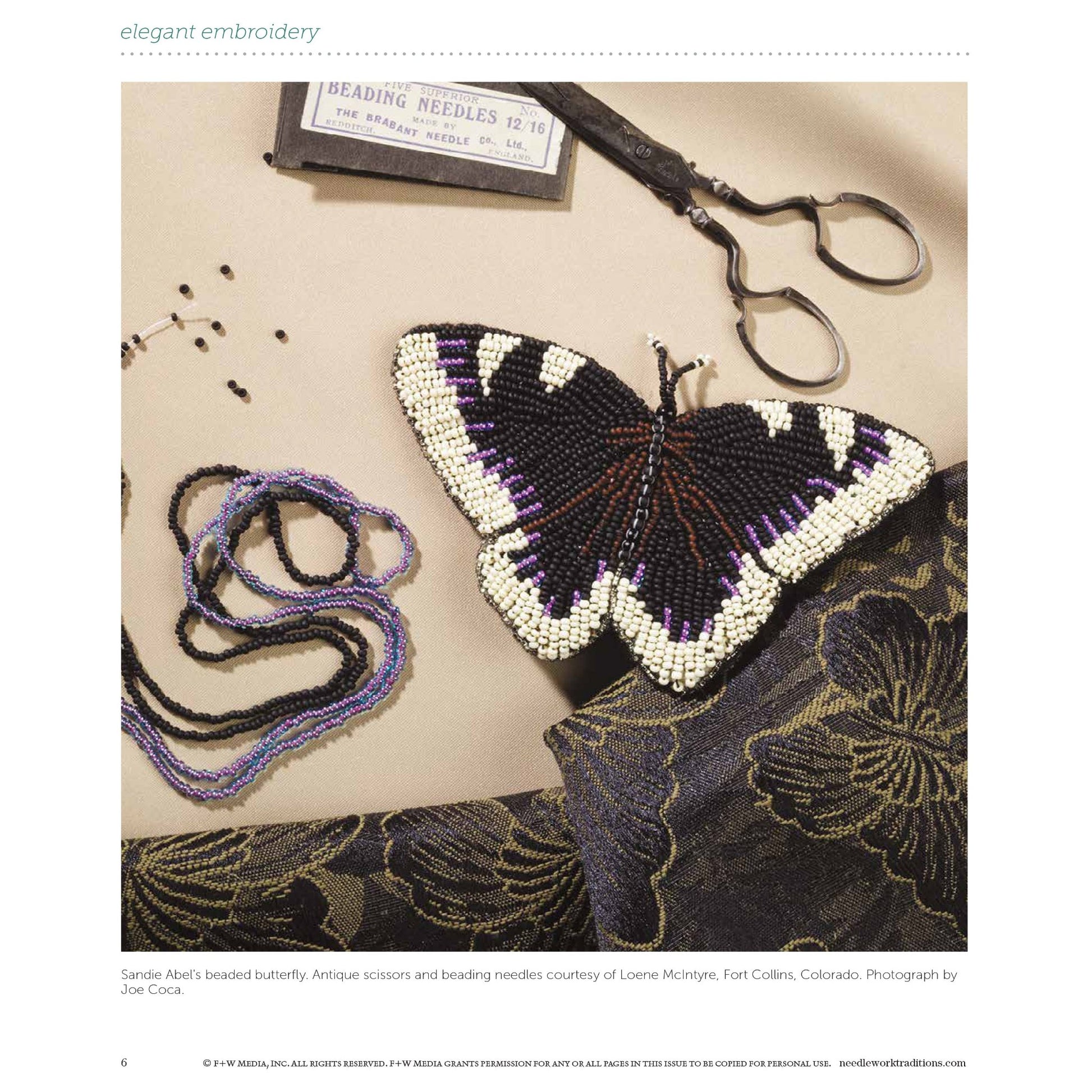 An intricately beaded butterfly in black, white, and purple sits on a richly patterned fabric, sourced from the "Elegant Embroidery with 5 Beautiful Projects to Stitch" eBook printed copy by Long Thread Media. Nearby are scissors and a small box of beading needles. Various strands of purple and black beads are also arranged around the butterfly.