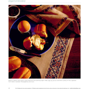 A table setting includes an embroidered linen placemat and napkin, featuring the intricate bands of Spanish stitch embroidery from **Long Thread Media’s "Elegant Embroidery with 5 Beautiful Projects to Stitch" eBook printed copy**, a blue plate with buttered bread rolls, an egg in a ceramic holder, a jam jar, and a knife.