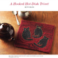 A teapot and spoon sit next to a hooked trivet adorned with three black cats on a textured red background, featuring simple, bold shapes that create a cozy and charming design—perfectly capturing the essence of the PieceWork Presents: Literary-Inspired Projects to Knit, Crochet, Hook, and Cross-Stitch eBook by Long Thread Media.