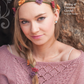 A person wearing a pink knit sweater and a headband featuring a variety of colorful knit flowers poses in front of a textured stone wall. The text reads, "String of Flowers Circlet from the Magical Knitted Costume Collection by Long Thread Media.