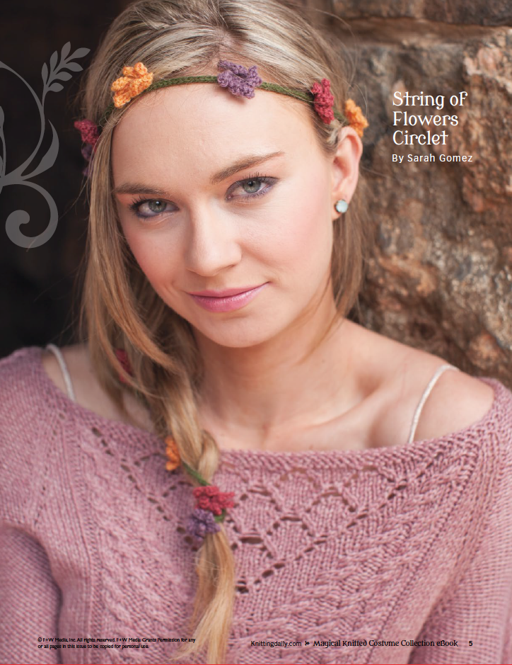 A person wearing a pink knit sweater and a headband featuring a variety of colorful knit flowers poses in front of a textured stone wall. The text reads, "String of Flowers Circlet from the Magical Knitted Costume Collection by Long Thread Media.