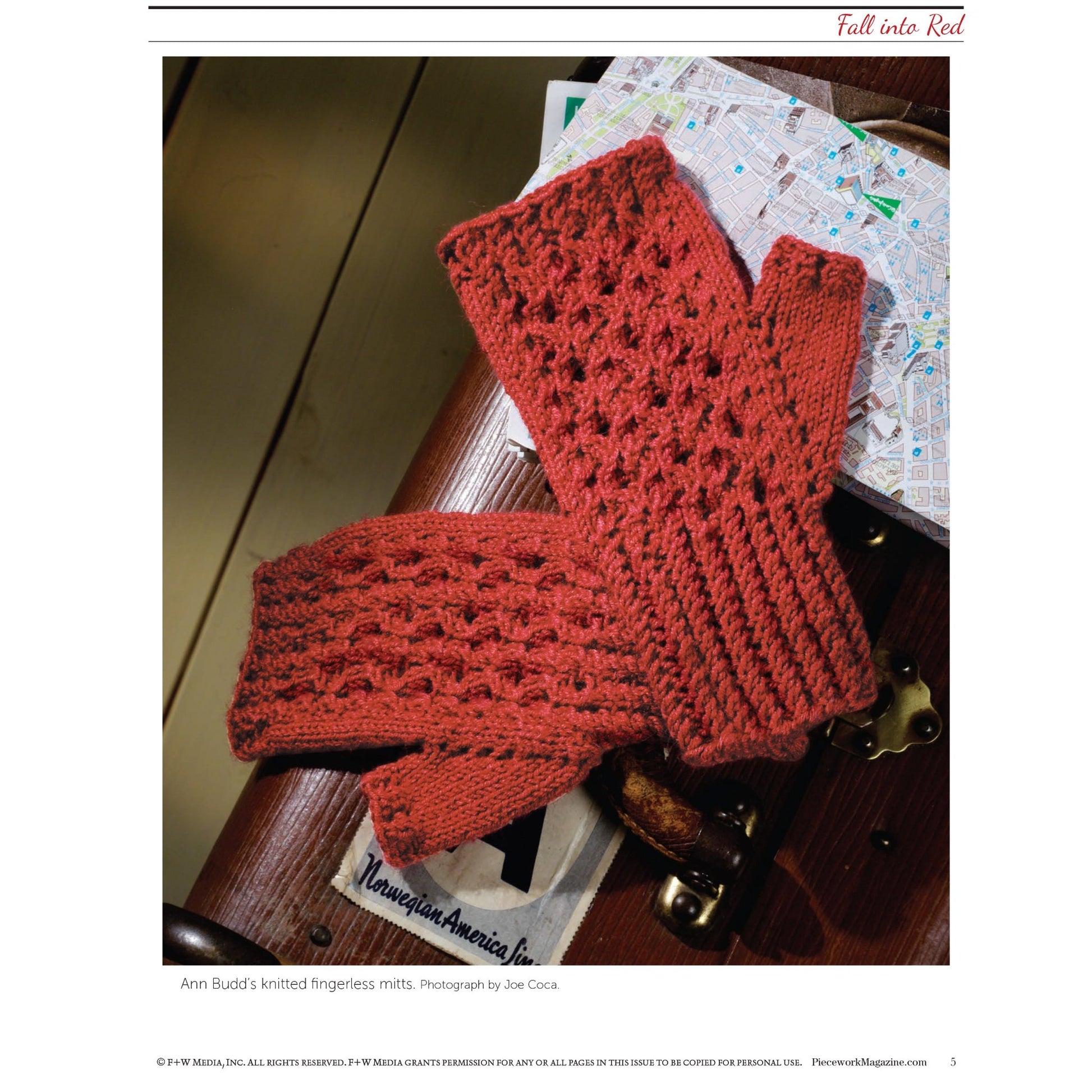 Displayed on a wooden surface is a pair of knitted, red fingerless mitts featuring an intricate lace pattern. In the background, there's a portion of a map and a small leather suitcase. The title "Fall Into Red: 4 Knitting Projects for the Season – eBook printed copy" from Long Thread Media is written at the top.