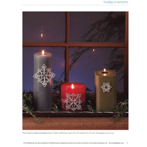 Three candles of various sizes and colors—gray, red, and green—are adorned with exquisite tatted snowflake ornaments crafted using the "PieceWork Holiday Ornaments: 5 Projects to Tat, Embroider, and Knit" eBook by Long Thread Media. Arranged on a windowsill amid lush green foliage, they create a festive holiday ambiance.