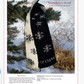 A knitted scarf, included in the "Winter Games: 4 Projects to Knit & More" eBook printed copy by Long Thread Media, is displayed hanging on a pine branch. The design features snowflake patterns with one side black featuring white snowflakes and the year “2014,” while the reverse side is white with black snowflakes, all set against a snowy landscape backdrop.