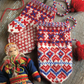 A copy of "Knitting the North: 4 Traditional Mitten Patterns from Northern Europe" by Long Thread Media is displayed on a wooden surface, showcasing colorful knitted children's mittens featuring traditional Sámi and Nordic designs in red, blue, and white. A doll in traditional Sámi attire sits beside the mittens, accompanied by yarn balls.