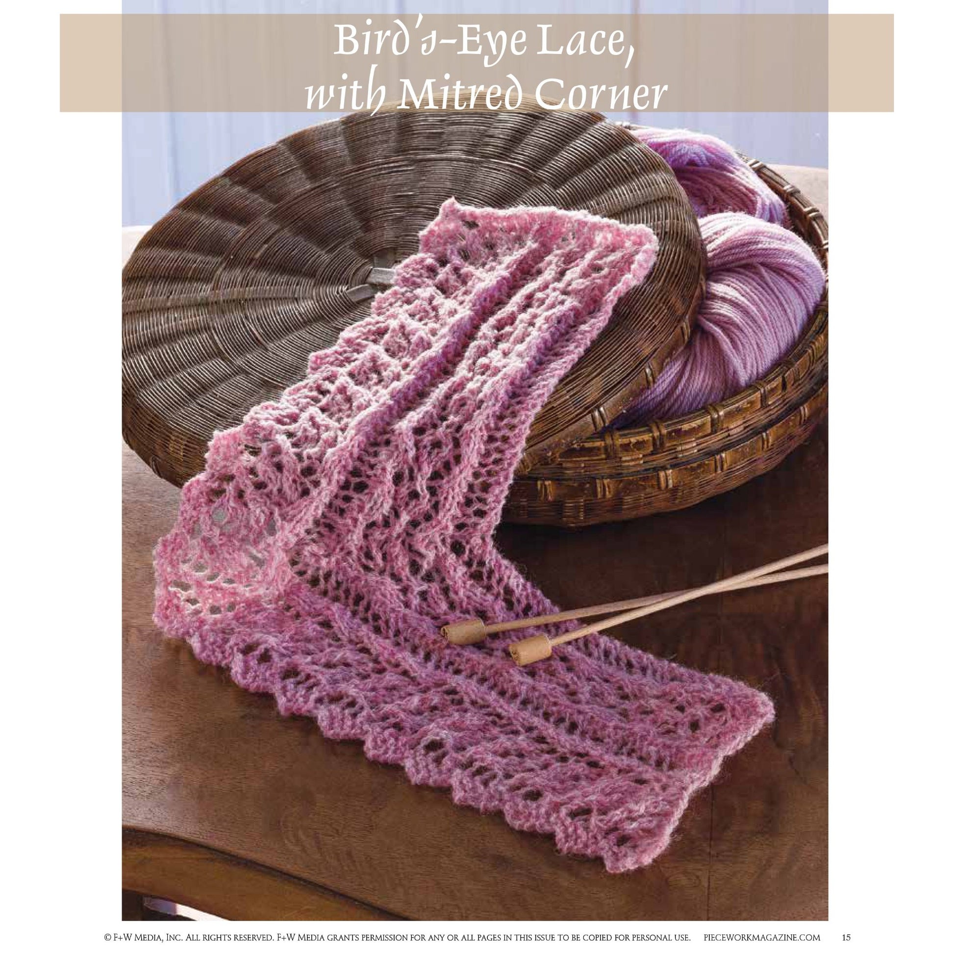 A delicate, pink lace piece knitted from the patterns in "Victorian Lace: 7 Timeless Projects to Knit – eBook printed copy" by Long Thread Media, is displayed partially folded on a wicker basket filled with purple yarn. Beside the lace, knitting needles lie elegantly. The backdrop features a wooden table bathed in soft, diffused light from a window.