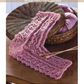 A knitted pink lace pattern named "Bird's-Eye Lace, with Mitred Corner" is elegantly spread over a tabletop, accompanied by a basket brimming with yarn. This design is part of the collection found in the eBook printed copy of "Victorian Lace: 7 Timeless Projects to Knit" by Long Thread Media.