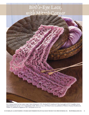 A knitted pink lace pattern named "Bird's-Eye Lace, with Mitred Corner" is elegantly spread over a tabletop, accompanied by a basket brimming with yarn. This design is part of the collection found in the eBook printed copy of "Victorian Lace: 7 Timeless Projects to Knit" by Long Thread Media.