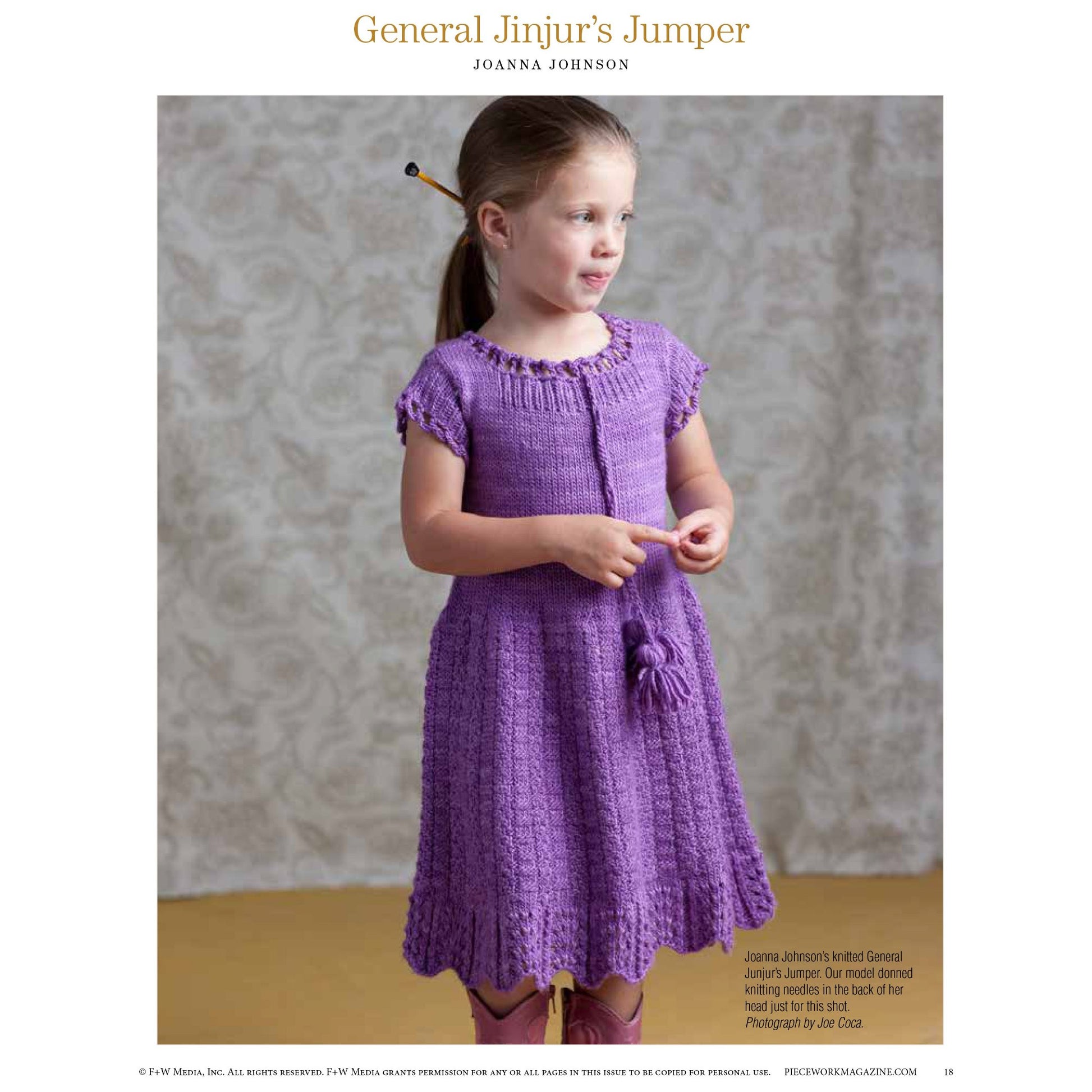 A young girl stands in a room with a patterned gray wall, wearing the "General Jinjur’s Jumper" from Joanna Johnson's **Victorian Times: 8 Projects to Knit** – an eBook published by Long Thread Media. She is adorned in a purple knitted dress with a flower detail and matching shoes. Clutched in her hand is a plush rabbit, and her hair is styled into pigtails.