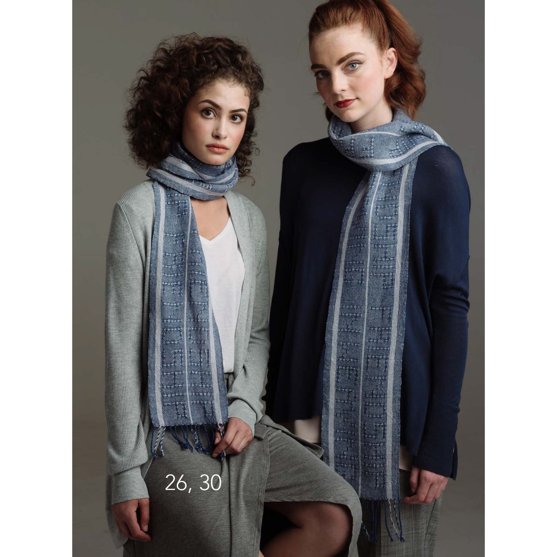 Two women stand closely together, both wearing long, light blue scarves from the Loom Theory: Eight & Over Eight Scarf Collection Shawl Collection by Long Thread Media. The woman on the left is dressed in a light grey cardigan over a white top and grey pants. The woman on the right wears a dark blue sweater and grey pants.