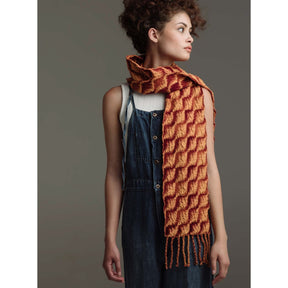 A person with curly hair wearing a sleeveless white top and blue overalls looks to the side. Draped around their neck is a chunky scarf from Long Thread Media's Loom Theory: Eight & Over Eight Scarf Collection, featuring vibrant orange and yellow woven patterns, with one end elegantly hanging down the front. The background is a neutral gray.