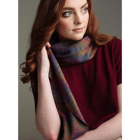 A woman with long, wavy red hair wears a dark maroon top and an elegant scarf from the Loom Theory: Eight & Over Eight Scarf Collection Shawl Collection by Long Thread Media. She is looking slightly to the side with a thoughtful expression against a plain gray background.