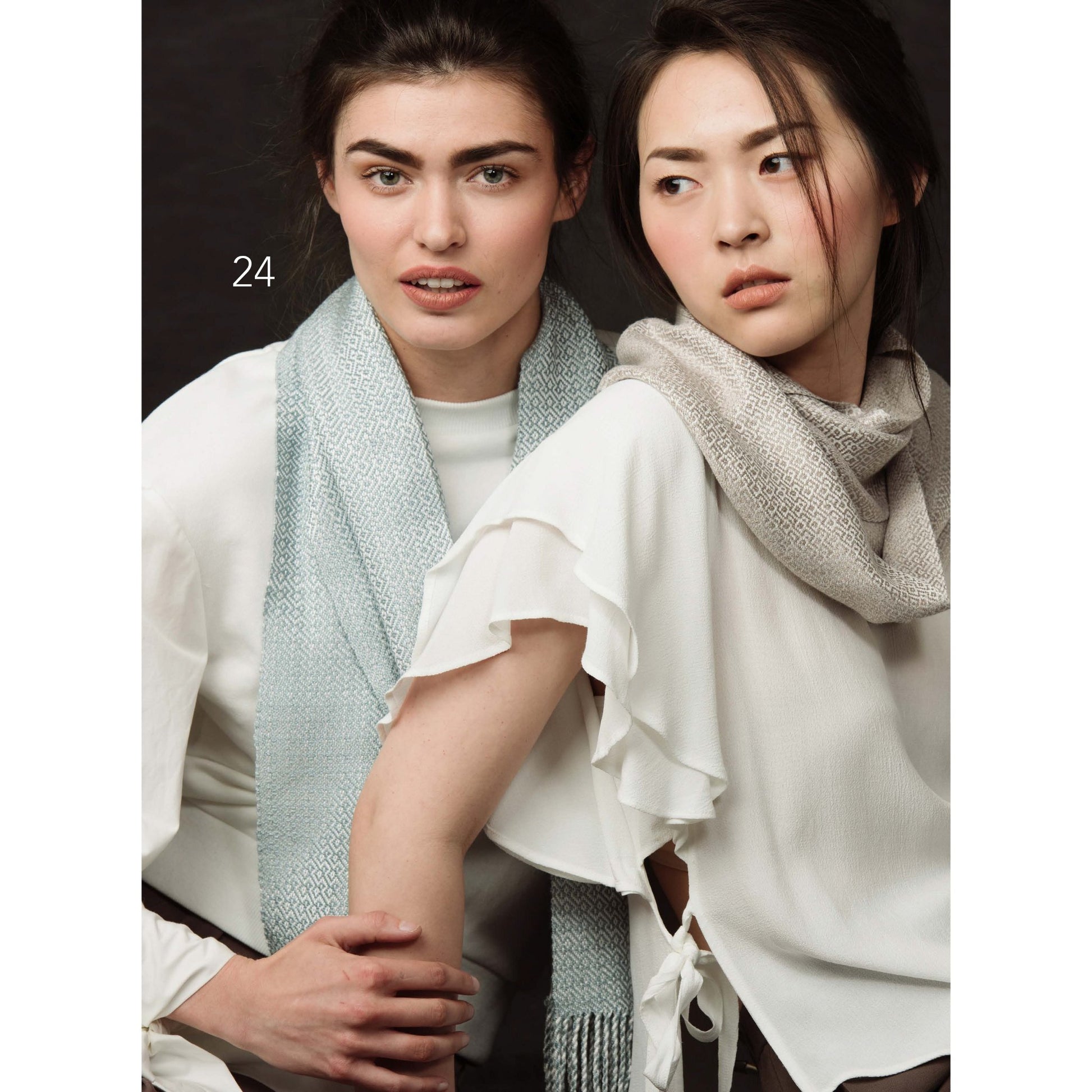 Two models stand against a dark background, each showcasing accessories from Long Thread Media's Handwoven Loom Theory: Four Shaft Scarf Collection. Both are dressed in white tops featuring ruffled details, with the model on the left gazing forward adorned in a light blue scarf, and the model on the right looking to the side with a grey scarf draped loosely.