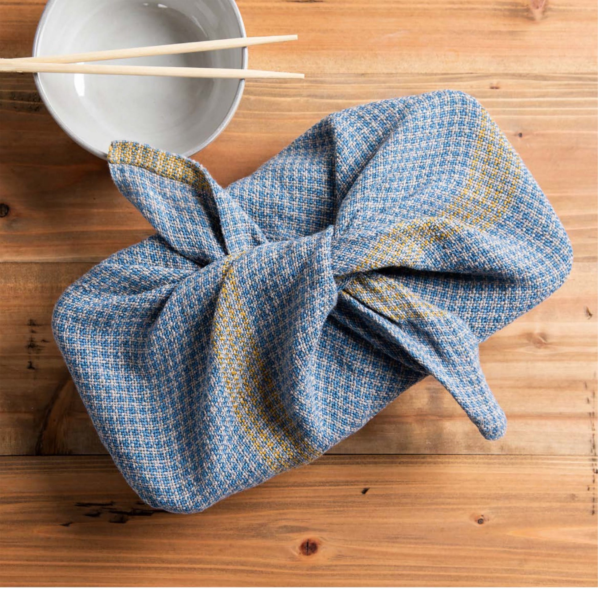 A copy of "Little Looms Presents: 10 Favorite Rigid Heddle Projects - eBook Printed Copy" by Long Thread Media, wrapped in blue fabric with a yellow pattern and tied at the top in a neat knot, sits on a wooden surface. Beside it, a pair of bamboo chopsticks rests next to a small white bowl.