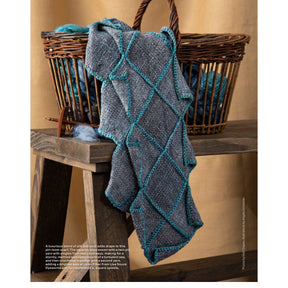 A textured, blue-gray woolen scarf with a teal grid pattern is draped over the edge of a wooden basket filled with yarn. The basket, sitting on a wooden table against a beige backdrop, showcases one of the intricate designs featured in "10 Favorite Pin-Loom Projects" by Long Thread Media.