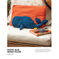 A blue woven whale pillow rests on an orange cushion, placed on an outdoor wicker chair. Nearby, a copy of "10 Favorite Pin-Loom Projects" by Long Thread Media, along with glasses and a small toy, add to the scene. The setting suggests a cozy, nautical-themed decor. Text includes the title "Mystic Blue Whale Pillow" and a description.