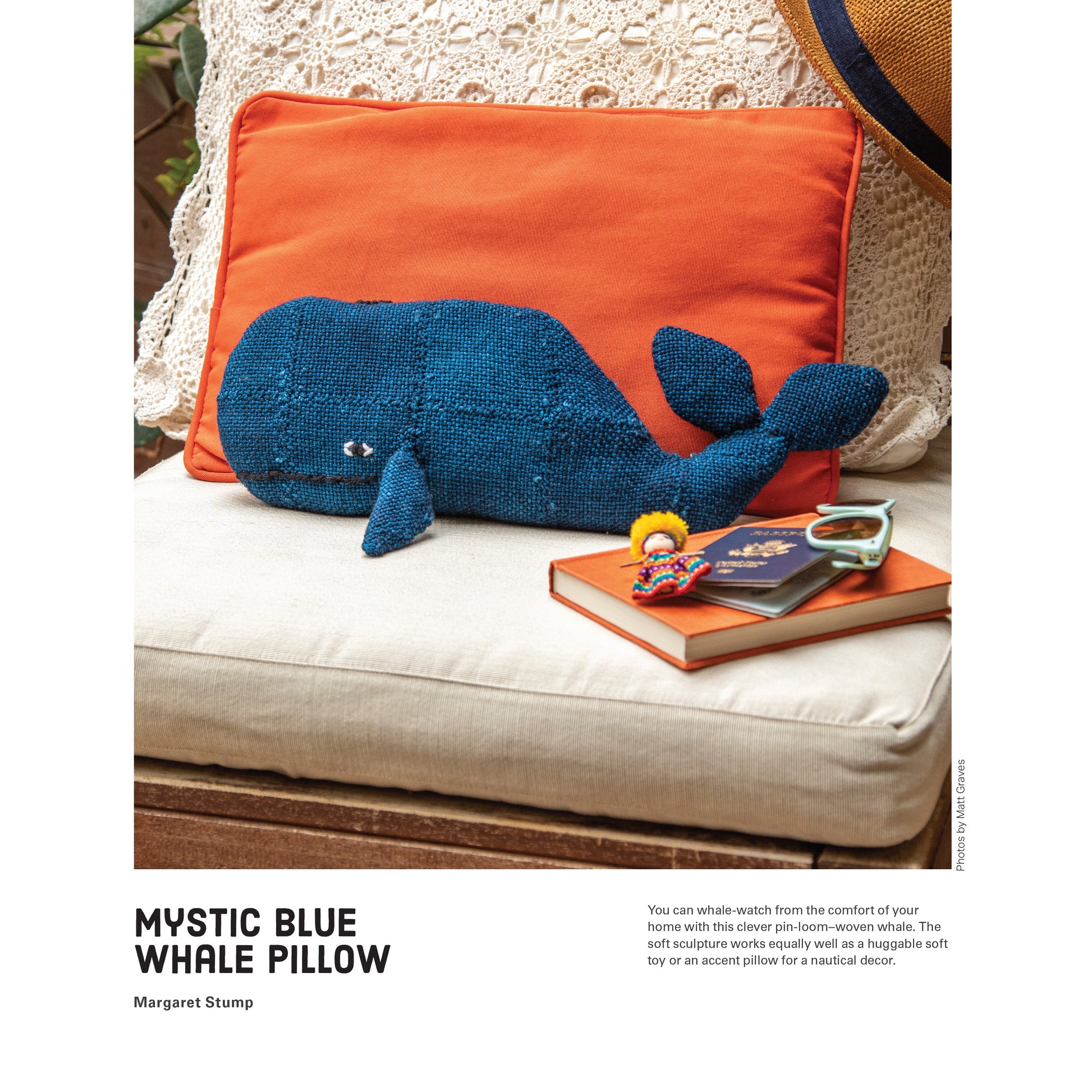 A blue woven whale pillow rests on an orange cushion, placed on an outdoor wicker chair. Nearby, a copy of "10 Favorite Pin-Loom Projects" by Long Thread Media, along with glasses and a small toy, add to the scene. The setting suggests a cozy, nautical-themed decor. Text includes the title "Mystic Blue Whale Pillow" and a description.