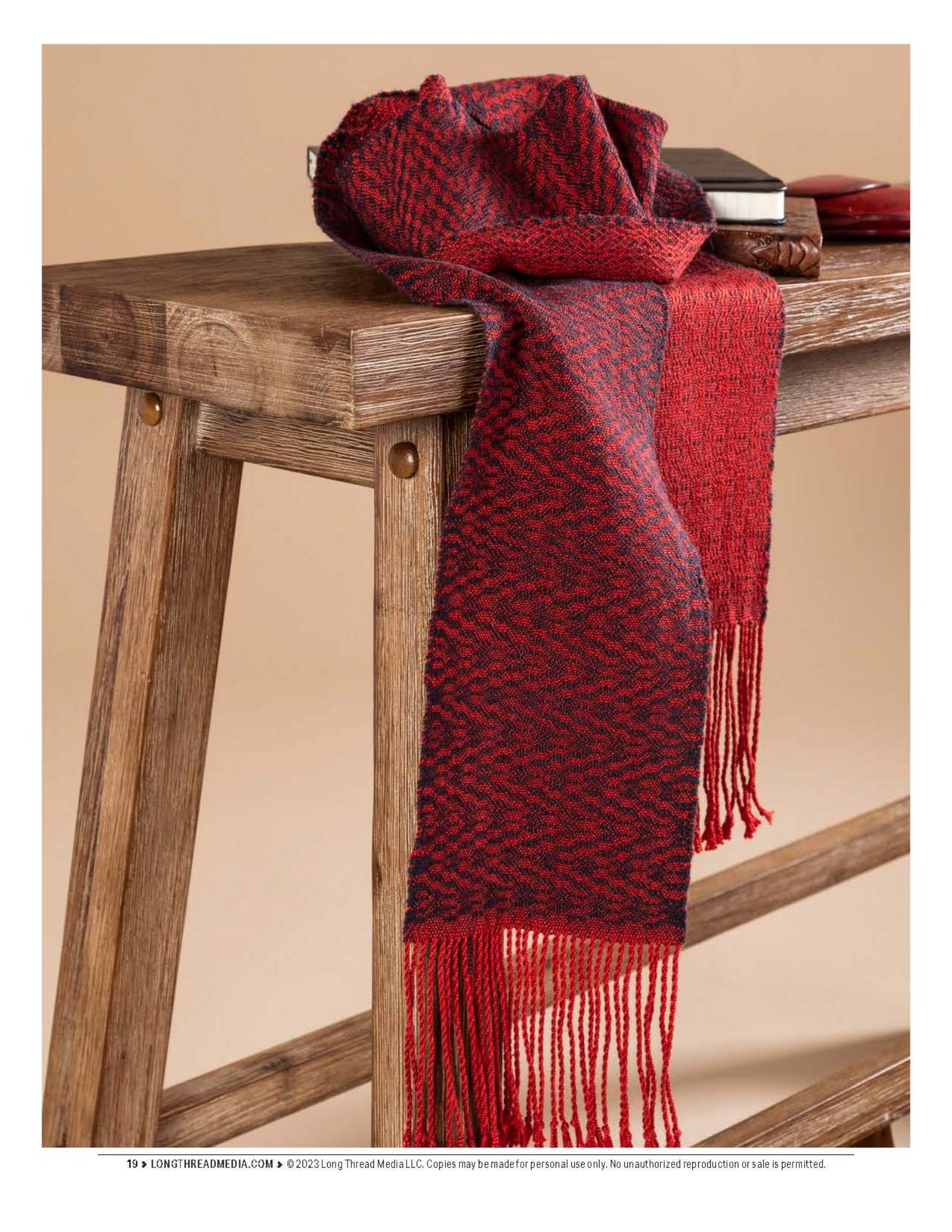 A copy of "Sensational Silk: Weave, Knit, Crochet, and Embroider with One of Nature’s Luxuries" by Long Thread Media is elegantly displayed over a rustic wooden table. The table seems to be crafted from reclaimed wood with visible grain and knots, contributing to a warm and cozy atmosphere.