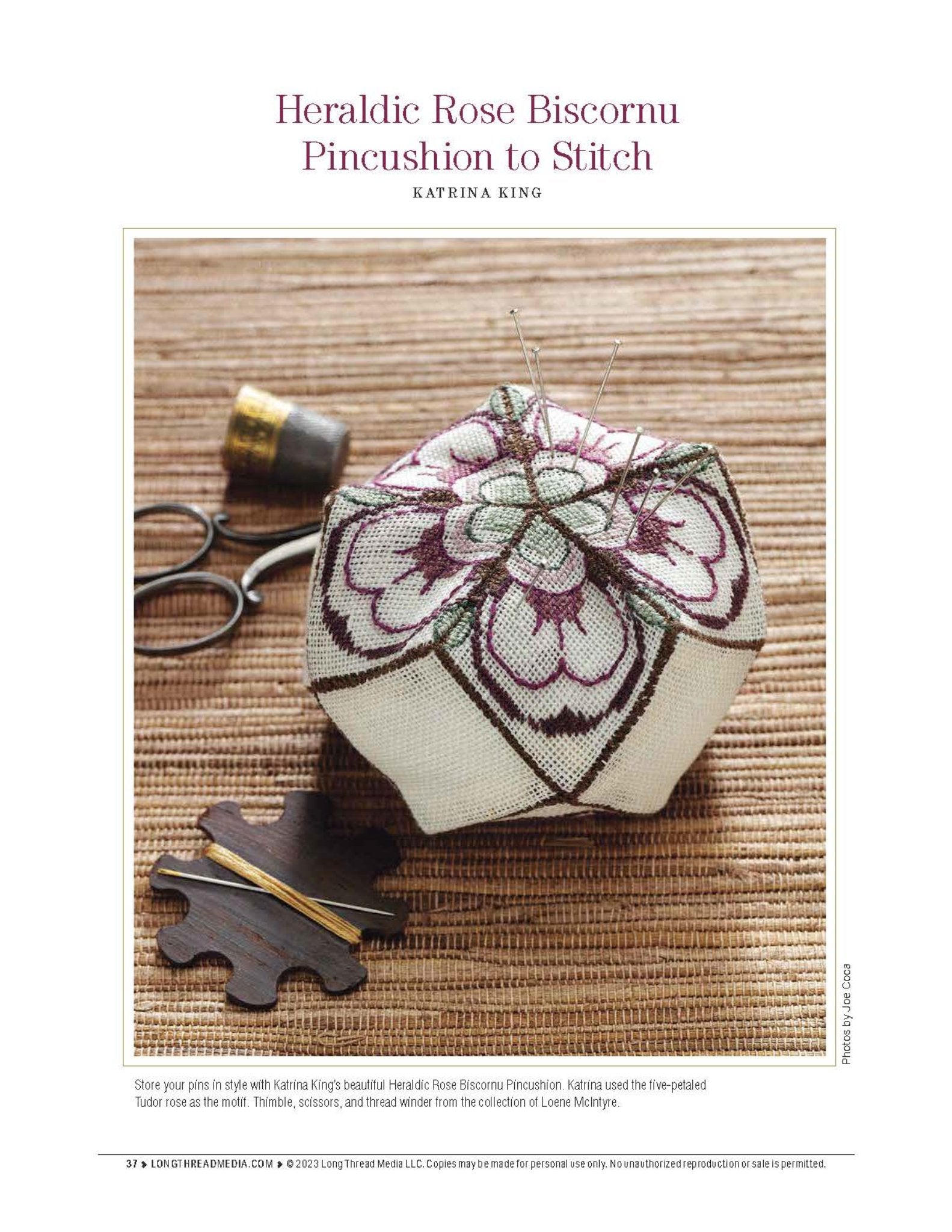 A detailed embroidery pincushion is featured with a pattern called "Heraldic Rose Biscornu," displaying a rose design in shades of pink and green on a white background. Accompanying the pincushion are scissors and a wooden thread holder, all placed on a textured surface. The scene exemplifies the beauty of silk projects showcased in "Sensational Silk: Weave, Knit, Crochet, and Embroider with One of Nature’s Luxuries," an eBook printed copy by Long Thread Media.