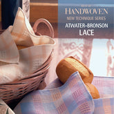 In a cozy breakfast scene, a basket of bread rolls sits beside pastel plaid napkins. In the background, the book "Best of Handwoven New Technique Series: Atwater-Bronson Lace eBook" by Long Thread Media is visible, suggesting intricate weaving techniques and delicate lace patterns.