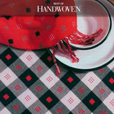 A table setting features a woven textile in red and black with geometric patterns beneath a white plate accented by a black rim. A fringed red cloth adorned with square designs elegantly drapes over the plate, capturing the essence of Long Thread Media's "Best of Handwoven, Top Ten Table Runners on Eight Shafts" eBook (Printed version). The title "Best of Handwoven" is prominently displayed at the top of the image.