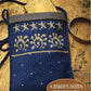 A small, blue handwoven bag adorned with beige star and sun patterns is showcased on a wooden surface, with a gold card partially visible at the top. The caption reads, "Best of Handwoven by Long Thread Media: A Baker's Dozen – 13 Bags to Weave.