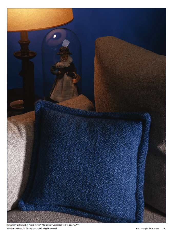 A blue textured pillow with intricate weaving designs sits on a beige couch near a lamp. In the background, a glass dome displays a small traditionally dressed figurine. The scene has a cozy, vintage feel and features Long Thread Media's "Best of Handwoven Technique Series: Twills on Four Shafts" eBook print edition.