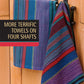 Two vibrant woven towels hang on hooks against a wooden backdrop. One towel showcases stripes in blue, green, and purple hues, while the other exhibits red, black, and blue stripes. Text reads "Best of Handwoven, More Terrific Towels on Four Shafts" by Long Thread Media.