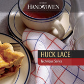 A tea set on a table showcases a white and blue teacup filled with tea, accompanied by a white teapot and a plate holding a slice of pie. A checkered cloth enhances the setting, while "Best of Handwoven Technique Series: Huck Lace," an eBook by Long Thread Media, highlighting exquisite weaving methods, is also on display.