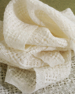 A soft, ivory-colored gauze fabric is loosely folded and layered, showcasing its delicate, open-weave texture. The backdrop of Long Thread Media's Handwoven's Master Weaver Collection: Favorite Projects and Lessons from Madelyn van der Hoogt eBook Printed Copy provides a subtle greenish hue, adding contrast to the light fabric.