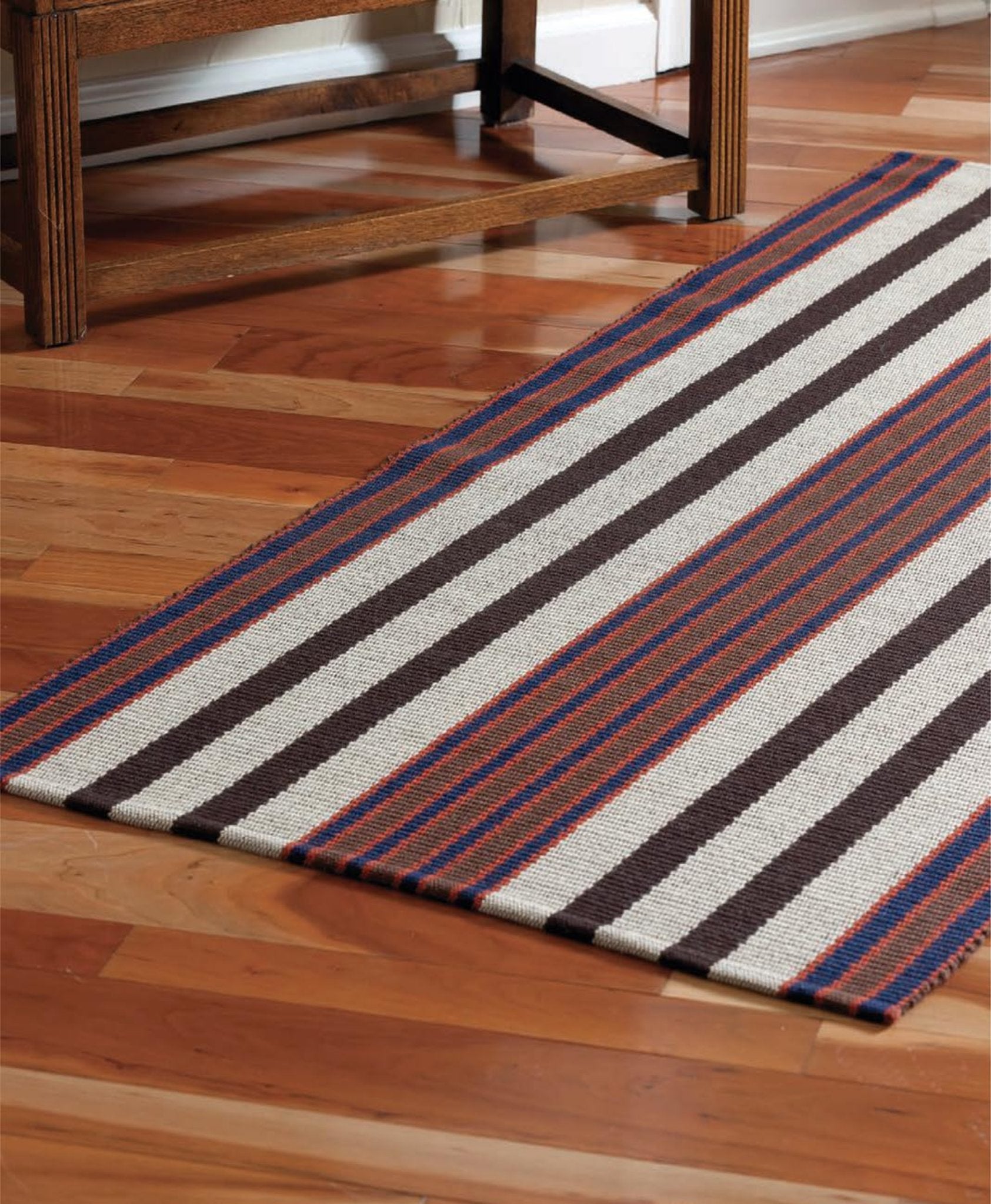 A rectangular rug featuring alternating stripes of brown, white, red, and blue lies on a polished wooden floor next to wooden furniture. The colorful contrast added by the rug enhances the aesthetic appeal, creating an inviting atmosphere reminiscent of Handwoven's Master Weaver Collection: Favorite Projects and Lessons from Tom Knisely - eBook Printed Copy by Long Thread Media.