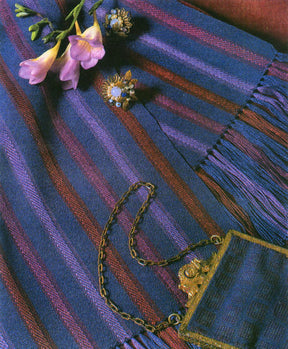 A handwoven cloth from the Master Weaver Collection: Favorite Projects and Lessons from Sharon Alderman eBook Printed Copy by Long Thread Media is laid out. On it rests a small ornate purse with a metallic clasp and chain, two delicate flower-shaped brooches, and a sprig of pink and yellow flowers. The cloth features fringed edges in shades of blue and purple.