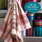 A dish towel featuring a red and white woven design hangs on a metal surface, with a collection of colorful stacked jars behind it. The text reads, "Long Thread Media Best of Handwoven: Top Ten Towels on Four Shafts (Printed Version).